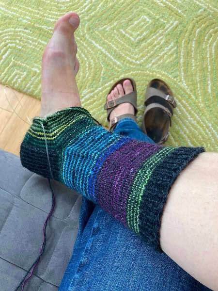 striped sock progress, shown modelled on foot, gusset decreases in progress. model is wearing jeans and sitting on a grey couch with a green rug