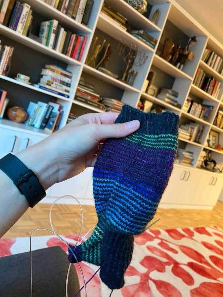 striped sock in progress, held up in a living room in front of white bookshelves which also hold other various art and sculpture.