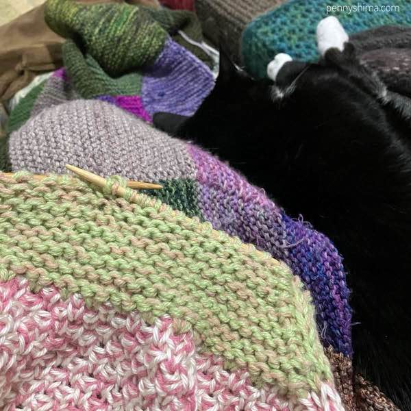 blanket in progress, pinks & green visible laid next to a tuxedo cat who is asleep