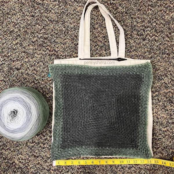 crochet square in grey-greens in progress, shown laid flat on top of a cotton tote bag. the cake of yarn is to the left. a yellow tape measure is along the bottom of the project.