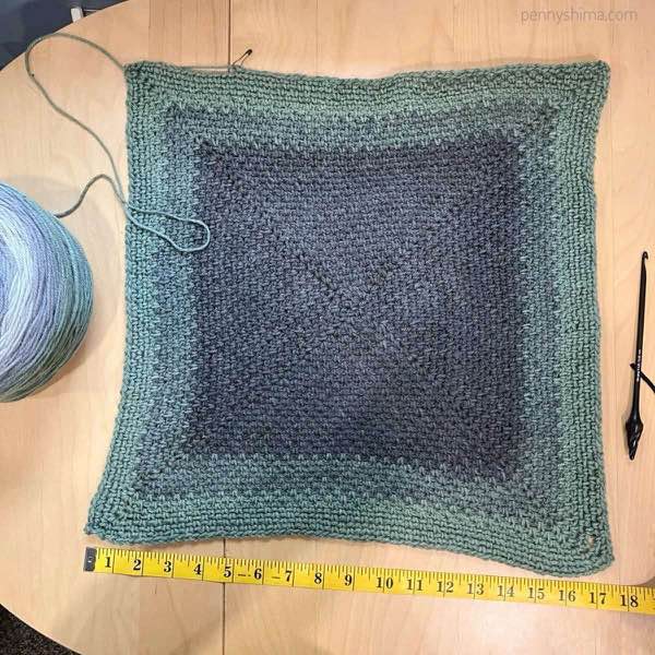 crochet square in grey-greens in progress, shown laid flat on a wooden surface