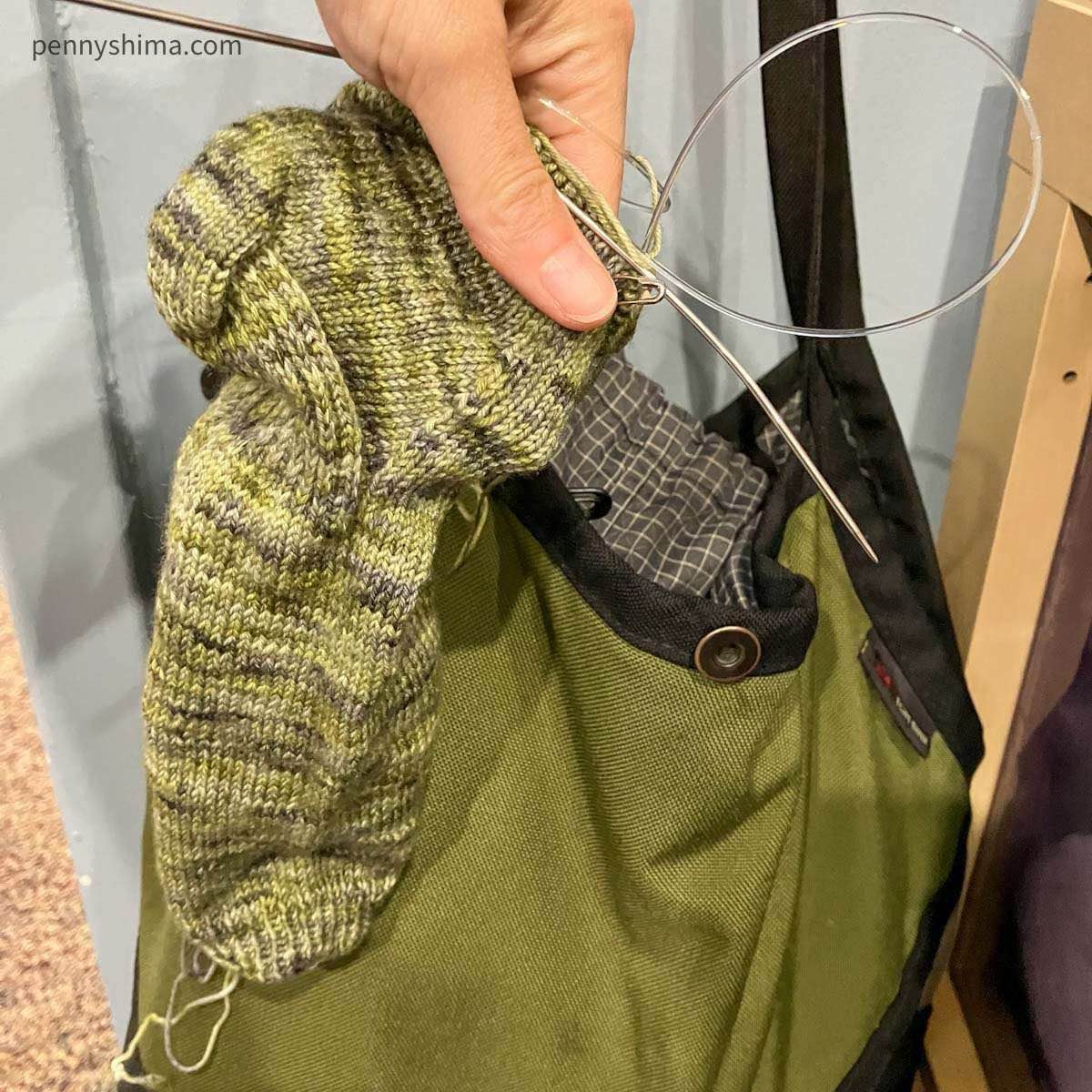 holding variegated green socks in progress in front of an olive green tom bihn swift that is hanging on a hook.