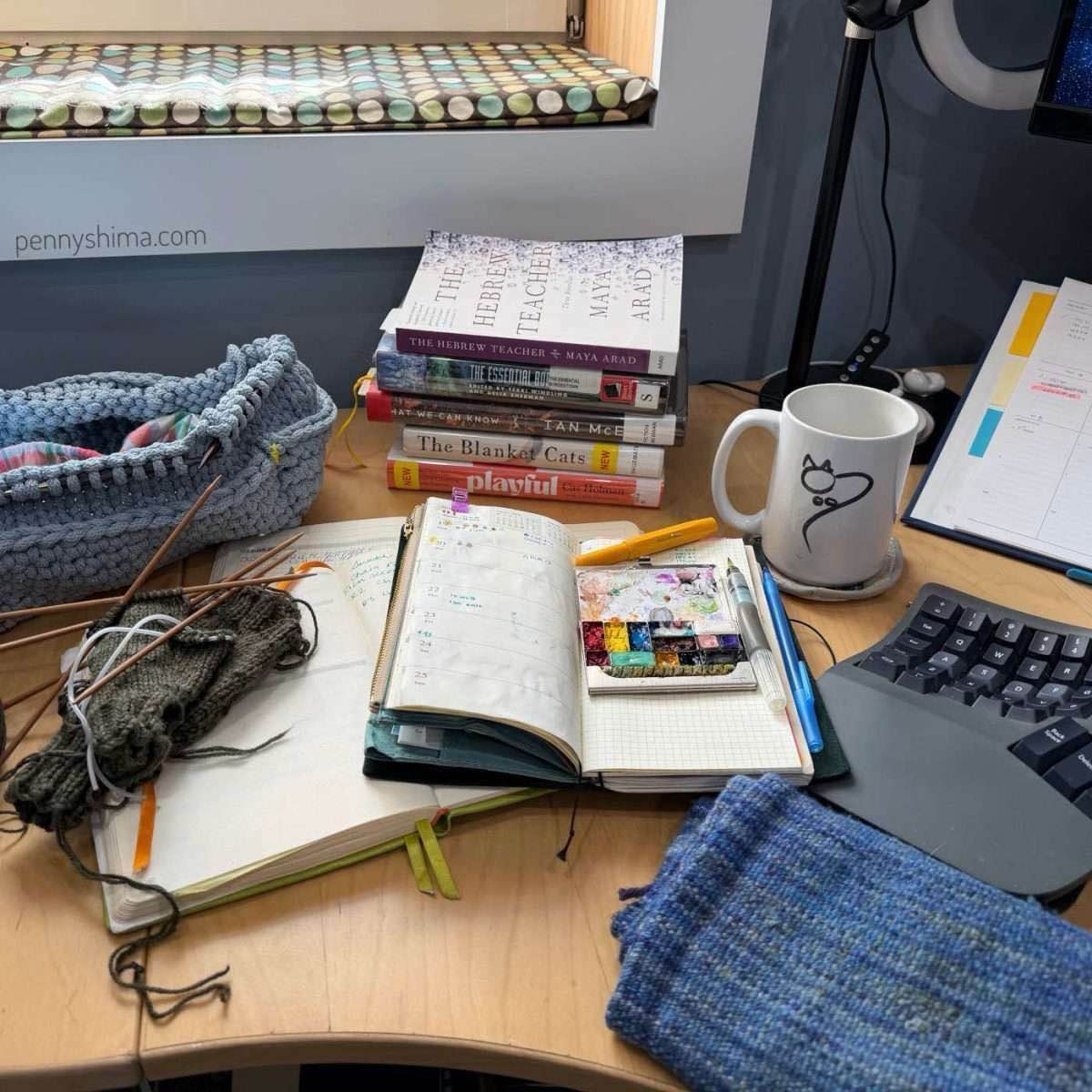 A Tuesday morning desk scene with a nearly empty coffee mug, part of an open planner visible, several notebooks, and a stack of five library books. Two knitting projects are visible in the workspace.
